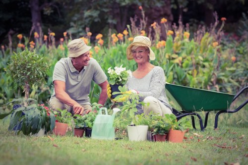 The image depicts a mature couple seated in a vibrant garden during daylight, surrounded by various potted plants and garden tools. The woman is holding a white flower and wearing a light-colored top, a wide-brimmed straw hat, and glasses, smiling warmly. The man, dressed in a casual grey T-shirt and also wearing a straw hat, appears engaged and cheerful. They are seated on the grass amidst a well-maintained garden with neat soil beds, lush green grass, and a backdrop of flowering plants and trees. To their side, there is a green wheelbarrow, a watering can, and several small pots with young plants, indicating active gardening and plant care. The garden environment is bright with natural sunlight, suggesting a calm, pleasant outdoor space suitable for gardening activities typical of Hampstead Heath. This scene naturally aligns with gardening services offered by landscapershampsteadheath.co.uk, emphasizing planting, lawn maintenance, and garden care.