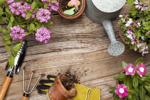 The image depicts a wooden outdoor garden table with gardening tools and supplies arranged on its surface. Visible elements include a small metal garden fork, a garden rake, a pair of gardening gloves in black and yellow, and a ceramic flower pot with soil and small colorful plant labels. Surrounding the tools are various flower pots filled with vibrant pink and purple flowering plants, with lush green leaves, suggesting they are in bloom. A grey watering can with a spout and drainage holes is positioned on the table, indicating garden watering activities. The weather appears to be bright and sunny, with natural light illuminating the scene, typical of a well-maintained outdoor garden space in Hampstead Heath. This setting emphasizes gardening and outdoor maintenance tasks, aligning with landscaping services focused on planting, flower bed care, and garden upkeep, reflective of a typical landscaped backyard or front garden environment in London.