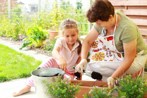 A woman and a young girl are engaged in gardening activities in a well-maintained outdoor space. The woman is wearing a floral apron and beige trousers, while the girl is dressed in a light pink top with pink and white striped pants, both working together with small garden tools. They are tending to a rectangular raised flower bed filled with dark soil, surrounded by lush, green plants and vibrant blooming flowers. In the foreground, there is a galvanized metal bucket and potted plants, with the garden extending into a grassy lawn area. The background shows a garden fence, potted plants, and a green hedge, indicating a tidy, landscaped garden typical of properties in Hampstead Heath. The setting appears to be a bright and mild day, ideal for outdoor gardening, with natural light illuminating the scene. This image reflects outdoor gardening and landscaping work suitable for gardening service providers like Landscapers Hampstead Heath, highlighting careful plant cultivation and garden maintenance in a residential garden environment near Hampstead, London postcode NW3.
