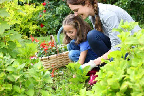 In a lush outdoor garden, a woman and a young girl are engaged in gardening activities amidst dense foliage and flowering plants. The woman, dressed in a light-colored top and dark jeans, is crouching down close to the ground, assisting the girl, who is wearing a blue t-shirt and jeans, with planting or tending to the plants. The garden features a well-maintained lawn with dense, green grass in the foreground and various shrubs and flowering bushes, including some with small red blossoms, surrounding the scene. In the background, there are taller trees providing shade and natural backdrop, while a light garden tool with a curved handle is partially visible near the side, hinting at outdoor maintenance. The scene is set in natural daylight, likely on a partly cloudy day, which highlights the vibrant green tones of the plants and the natural textures of soil, grass, and foliage. This image reflects a typical garden scene that a professional landscaping service like Landscapers Hampstead Heath might support, emphasizing the area's lush, well-kept outdoor spaces around Hampstead Heath postcode area, suitable for gardening and planting services.
