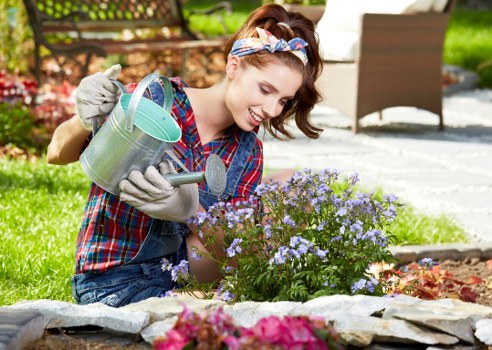 A young woman with light skin, wearing a plaid shirt, jeans, gardening gloves, and a colorful headband, is kneeling on a stone-edged flower bed in a residential garden. She is watering a cluster of purple and light blue flowering plants with a green metal watering can. The garden features a neatly maintained lawn, with a paved patio area in the background, partly shaded by trees and garden furniture. Surrounding the flower bed are various plants and shrubs, with a hedge visible further back, creating a lush, well-kept outdoor space typical of Hampstead Heath gardens. The scene appears to be on a sunny day with natural daylight illuminating the vibrant colours of the flowers and greenery, and the woman’s cheerful expression suggests enjoyment in gardening activities. This setting reflects typical gardening services such as planting, lawn care, and outdoor maintenance offered by Landscaping Hampstead Heath, aligning with the focus on landscaping and garden upkeep for residential properties in NW3 postcode area.