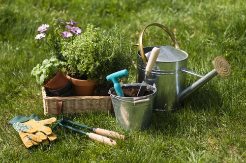 A well-maintained garden features a lush green lawn with evenly cut grass, bordered by various potted plants including flowering plants with pink and purple blooms, and green foliage in terracotta and plastic pots. There is a wooden garden bed containing more greenery, situated near the backdrop of a hedge line or shrubbery. In the foreground, gardening tools such as a pair of yellow gloves, a small hand trowel, and garden shears are placed on the grass, alongside a metal watering can with a wooden handle and a smaller watering can, indicating ongoing outdoor maintenance. The scene is set in natural daylight under overcast or partly cloudy weather, creating a soft, diffused light that highlights the rich textures and natural colours of the garden elements. This outdoor space exemplifies typical garden care and landscaping activities, supporting services offered by companies like landscapershampsteadheath.co.uk in the Hampstead Heath area, with an emphasis on lawn care, planting, and garden beautification.