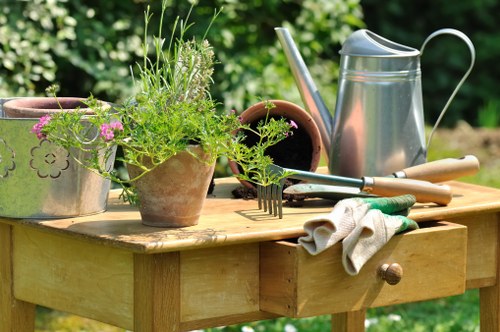 A wooden garden table set outdoors in a lush green garden with various gardening tools and plant pots. The table holds a metal watering can, a pair of gardening gloves, a small hand rake, and two terracotta pots, one filled with vibrant green foliage and delicate purple flowers. In the background, dense shrubbery and trees create a natural, serene environment, hinting at well-maintained garden spaces typical of properties near Hampstead Heath. The scene is illuminated by natural daylight, suggesting a bright, clear day suitable for outdoor gardening activities. This setup reflects a well-kept garden area suitable for professional gardening and landscaping services, such as fence repairs, to enhance outdoor space usability and aesthetics, aligning with local gardening service offerings in Hampstead.