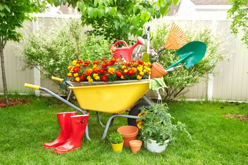 A brightly colored garden wheelbarrow with a yellow body and silver handles is positioned on a well-maintained grassy lawn in a residential outdoor space. The wheelbarrow is filled with vibrant red and orange flowers, adding a splash of natural colour to the scene. Surrounding the wheelbarrow are gardening tools and accessories, including a pair of red rubber boots leaning against it, a small yellow watering can, and several terracotta plant pots placed on the grass. In the background, lush green trees and shrubs provide a leafy backdrop, with a white fence delineating the garden boundary. The scene suggests a tidy and inviting garden environment suitable for planting, lawn care, and outdoor maintenance, characteristic of professional landscaping services in Hampstead Heath. The overall setting is illuminated by natural daylight, indicating a clear, pleasant weather condition, emphasizing the garden's vibrant and well-kept appearance. This outdoor space exemplifies a typical residential garden that can benefit from expert planting solutions and garden management offered by landscapers Hampstead Heath.