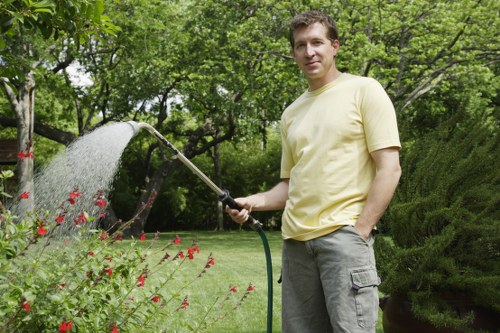 A man stands in a well-maintained garden in Hampstead Heath, holding a garden hose that is watering a bed of red flowering plants. The garden features a lush, green lawn with neatly edged flower beds, bordered by various shrubs and trees with dense foliage. In the background, tall trees and a hedge create a natural backdrop, with dappled sunlight filtering through the leaves, indicating a bright day with clear weather. The scene showcases a tidy outdoor space with vibrant plant life, suitable for gardening services focused on lawn care and garden maintenance within the Hampstead Heath area.