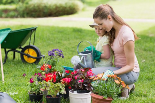 In a well-maintained garden, a woman and a young girl are engaging in planting activities among a variety of vibrant flowering plants in pots, including red, pink, and purple blooms, placed on the lush, green grass. The background features a neatly trimmed hedge and mature trees, with a lawn area extending into the distance, suggesting a spacious outdoor environment typical of a Hampstead Heath residential garden. To the left, a green and yellow garden wheelbarrow is partially visible, indicating ongoing gardening work. The sky appears clear, providing natural daylight that enhances the natural tones of the plants, wooden decking, and grass. The scene reflects a peaceful moment of outdoor gardening, supported by professional gardening services such as those offered by Landscapers Hampstead Heath, focused on garden maintenance, planting, and landscape improvements within the local area.
