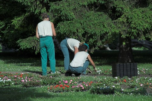 The image depicts three individuals working together in a well-maintained garden, with two of them actively engaged in planting or tending to the flower beds, while the third stands nearby observing. The garden features a lush, green lawn in the foreground, bordered by vibrant flower beds filled with small, colorful blooms, possibly annuals or perennials. Behind the group, there is a dense canopy of mature trees with abundant foliage, providing shade and a natural backdrop consistent with outdoor spaces in Hampstead Heath. The scene takes place in daylight under clear weather conditions, highlighting the healthy, vibrant plant life and the natural textures of the grass, soil, and branches. The garden appears to be part of a residential or community outdoor area, maintained with care, reflecting professional landscaping practices typical of local gardening services like Landscapers Hampstead Heath. This setting underscores a focus on environmentally conscious gardening and sustainable outdoor management, aligning with themes of recycling and sustainability featured on the webpage.