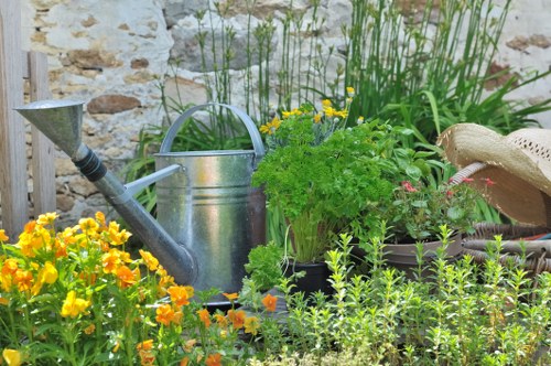 A small, vibrant garden scene featuring a variety of plants, including bright yellow and orange flowering plants in the foreground, complemented by lush green foliage and herbs in containers. The garden is bordered by a rustic stone wall, with taller grasses and plants growing behind the containers. An aged metal watering can and a straw hat rest among the plants, suggesting recent gardening activities. The ground surface appears to be a mix of soil and small paving stones, creating a practical and inviting outdoor space suitable for gardening and lawn care, typical of a residential Hampstead Heath area. The overall atmosphere is natural and well-maintained, with soft natural lighting indicating a mild, partly cloudy day, emphasizing the textures and colours of the plants and garden accessories. This scene supports gardening services such as planting, maintenance, and outdoor aesthetic improvements, aligning with the local garden environment in Hampstead Heath.