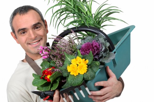 The image depicts a smiling man holding a teal plastic garden trowel with a small flowerpot containing a lush, green, tall plant with elongated leaves. In the foreground, a black plastic container is filled with a variety of flowering plants, including a yellow primula, purple ornamental cabbage, and small red flowers, arranged to create a colourful display. The background suggests an outdoor garden environment with natural light illuminating the scene, indicating a clear or partly cloudy day. The combination of garden tools, vibrant plants, and outdoor setting reflects professional gardening and landscaping work, typical of a landscaping service in Hampstead Heath. The scene highlights elements associated with flower bed creation and outdoor horticultural care, aligning with gardening services offered by Landscapers Hampstead Heath in the local area.