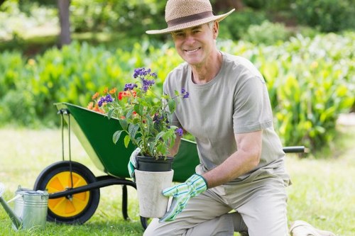 A gardener wearing a straw hat, grey t-shirt, and gardening gloves kneels on a well-maintained lawn in an outdoor space, holding a potted plant with purple, red, and pink flowers. Behind him, there is a green wheelbarrow with a yellow wheel, which appears to be used for garden tools or compost, on a lush, green lawn with a backdrop of dense greenery, including trees and shrubs. The area features a neatly edged flower bed or border, and the scene is lit by natural daylight, suggesting a clear and mild weather day. The garden environment indicates a landscaped yard typical of a Hampstead Heath residential area, emphasizing lawn care, planting, and outdoor maintenance, consistent with services offered by Landscapers Hampstead Heath.