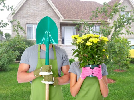 A person wearing a teal gardening helmet, grey t-shirt, and green gardening gloves is holding a yellow-handled shovel vertically in front of them on a well-maintained lawn. Next to them, another person in a grey t-shirt and pink gardening gloves is holding a large pink pot filled with blooming yellow flowers, obscuring their face. The background features a suburban garden with a neatly trimmed lawn, mature hedges, small trees, and a brick house with white-framed windows and a sloped roof. The scene is set outdoors on a cloudy day, with natural light illuminating the garden’s green grass, foliage, and vibrant flowers. This image demonstrates outdoor work related to gardening and planting, consistent with the services offered by Landscapers Hampstead Heath in the Hampstead Heath area, focusing on garden planting, lawn care, and landscape maintenance in a private residential setting.