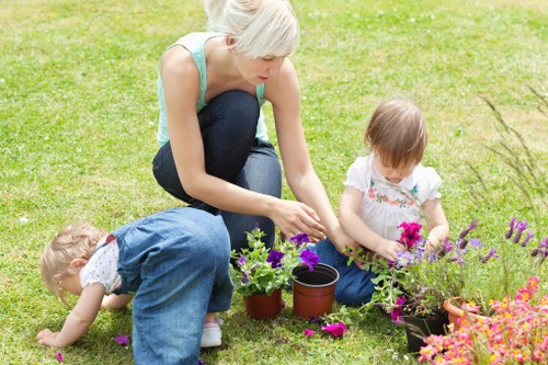 A woman with short blonde hair and light skin is kneeling on a well-maintained green lawn in a garden, surrounded by two young light-skinned children, one with curly blonde hair and the other with straight brown hair. The woman is tending to potted flowering plants, including pink, purple, and orange blooms, with some plants placed on the grass and others in black plastic pots. The garden features lush grass, a flower bed with a variety of colourful flowers in the background, and a backdrop of trees and shrubs. The scene appears to be outdoors on a bright, mild day, capturing a moment of outdoor gardening activity typical of landscaped yards that local gardening services, such as Hampstead Heath-based landscapers, might support by maintaining vibrant lawns and flower beds for residential outdoor spaces.