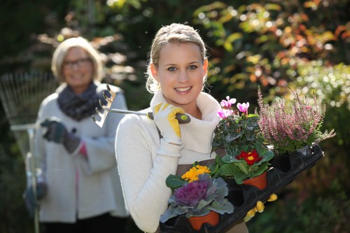 A young woman with blonde hair, wearing a white sweater and yellow gardening gloves, is standing outdoors in a well-maintained garden during daylight hours. She is smiling and holding a black gardening tray filled with colorful flowering plants, including pink, purple, yellow, and red blooms. Behind her, an older woman with glasses, also dressed in outdoor clothing, is slightly out of focus and holding garden tools, indicating a gardening activity in a lush, green garden with trees, shrubs, and a neatly trimmed hedge. The scene suggests a pleasant, sunny day suitable for gardening and outdoor landscaping, potentially part of a gardening service in Hampstead Heath or nearby areas, emphasizing professional outdoor maintenance and garden enhancement.